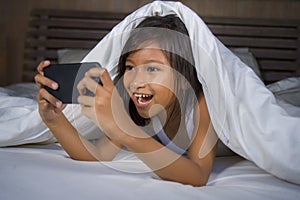 Happy and beautiful 7 years old child having fun playing internet game with mobile phone lying on bed cheerful and excited in