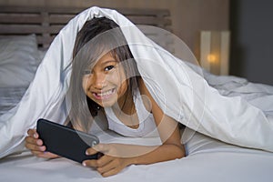 Happy and beautiful 7 years old child having fun playing internet game with mobile phone lying on bed cheerful and excited in