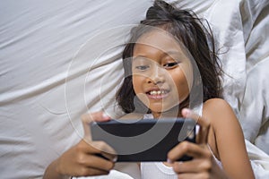 Happy and beautiful 7 years old child having fun playing internet game with mobile phone lying on bed cheerful and excited in