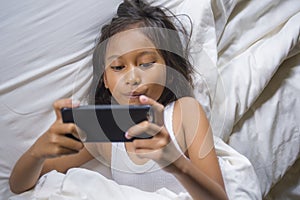 Happy and beautiful 7 years old child having fun playing internet game with mobile phone lying on bed cheerful and excited in