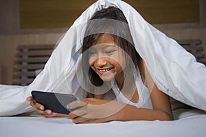 Happy and beautiful 7 years old child having fun playing internet game with mobile phone lying on bed cheerful and excited in kid