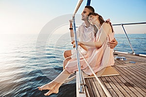 Happy beautiful adult couple sitting on side of yacht, watching at seaside and hugging while on vacation. Tan might fade