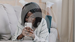 Happy beautiful african american girl lying on cozy sofa resting holding phone young woman chatting with friends online