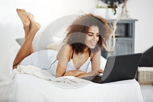 Happy beautiful african girl in sleepwear lying on bed at home smiling looking at laptop.