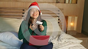 Happy and beautiful Asian American girl in Santa Christmas hat using credit card and laptop computer for online shopping