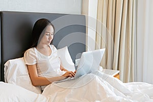 Happy beautiful Asian businesswoman working on a laptop while sitting on bed at home. Work from home. Technology and lifestyle