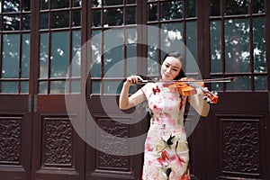 Happy beautiful Asian Chinese woman artist in traditional chi-pao cheongsam in a garden play musical instruments violin art