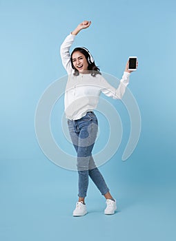 Happy beautiful Asian teen woman listening music on mobile phone wear headphones and dance on blue background