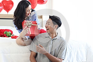 Happy beautiful Asian woman giving her boyfriend a red present gift box while sitting on white bed in bedroom, romantic lover