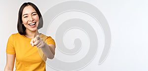 Happy beautiful asian woman laughing, pointing finger at camera and chuckle, smiling carefree, standing in yellow tshirt