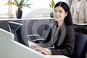 Happy beautiful Asian woman officer typing on desktop computer while sitting at office desk with blurred background of her busy