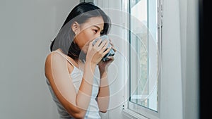 Happy beautiful Asian woman smiling and drinking a cup of coffee or tea near the window in bedroom.