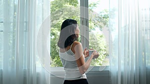 Happy beautiful Asian woman smiling and drinking a cup of coffee or tea near the window in bedroom. Young latin girl open curtains