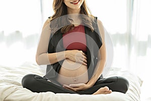 Happy Beautiful Attractive Pregnant Asian Woman sitting on bed smile and holding her big belly