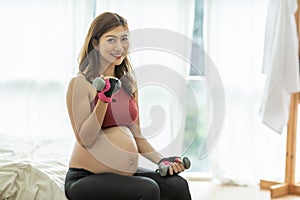 Happy Beautiful Attractive Pregnant Asian Woman standing near windows and workout exercising with dumbbell