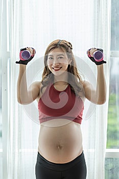 Happy Beautiful Attractive Pregnant Asian Woman standing near windows and workout exercising with dumbbell smile with big belly