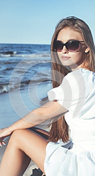 Happy, beautiful blonde woman is sitting on the ocean beach in a white summer dress and sunglasses