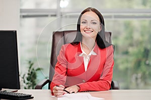 A young attractive Caucasian blond woman in a red business suit sits at a desk in a bright office. Portrait of a