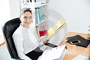 Happy beautiful businesswoman holding binders while sitting at the office