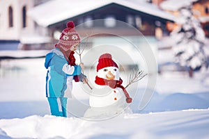 Happy beautiful child building snowman in garden, winter