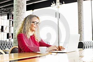 Happy beautiful curly hair blonde young female at coffee shop using laptop, smiling. Young attractive woman freelancer wearing gla