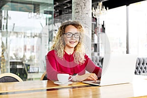 Happy beautiful curly hair blonde young female at coffee shop using laptop, smiling. Young attractive woman freelancer wearing gla