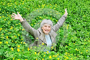 Happy beautiful elderly woman sitting arms outstretched on a glade of yellow flowers in spring.