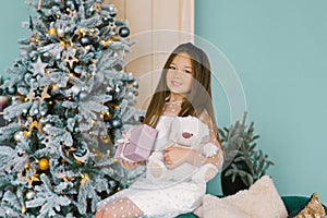 Happy beautiful girl holding a gift box and a stuffed bear toy and sitting near the Christmas tree on the sofa