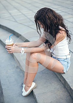 Happy beautiful girl sitting on stairs in park and drinking ice coffee