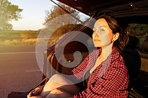 happy beautiful girl sitting in the trunk of car and enjoying sunset on the roadside. young woman in the car