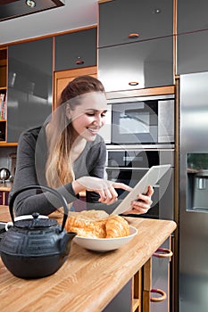 Happy beautiful girl touching her tablet in kitchen with croissants