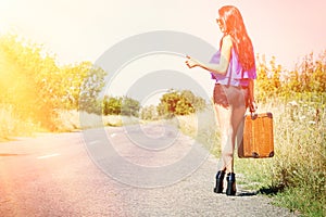 Happy beautiful girl traveler with a suitcase on the road, hitchhiking. The concept of travel, adventure, vacation, freedom.