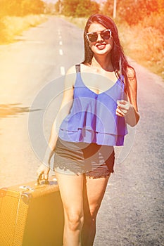 Happy beautiful girl traveler with a suitcase on the road, hitchhiking. The concept of travel, adventure, vacation, freedom.