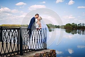 happy and beautiful groom and bride tender kiss at spring outdoors at the beach of the river under blue sky