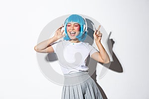 Happy beautiful japanese girl in blue wig listening music in headphones, dancing and singing along, standing over white