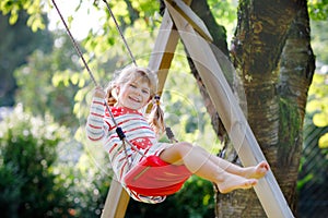 Happy beautiful little toddler girl having fun on swing in domestic garden. Cute healthy child swinging under blooming