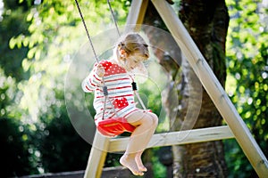 Happy beautiful little toddler girl having fun on swing in domestic garden. Cute healthy child swinging under blooming