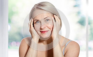 Happy beautiful middle-aged blonde woman shows off her perfectly well-groomed face and sitting relaxed near the window