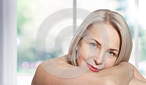 Happy beautiful middle-aged blonde woman shows off her perfectly well-groomed face and sitting relaxed near the window