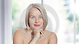 Happy beautiful middle-aged blonde woman shows off her perfectly well-groomed face and sitting relaxed near the window