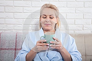 Happy beautiful middle aged woman drinking coffee on sofa at home