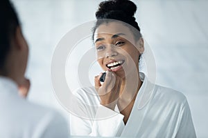 Happy beautiful millennial Black girl putting on lipstick, rouging lips