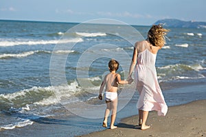 Happy beautiful mom and child walk far away along the sea beach, wind blowing