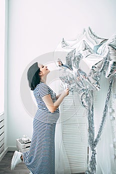 Happy beautiful pregnant caucasian woman with big belly in dress in the child room with balloons, future parrents