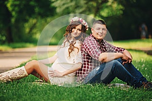 Happy beautiful pregnant woman with his husband sitting on the grass outdoors in the park on summer day. pregnant woman expecting