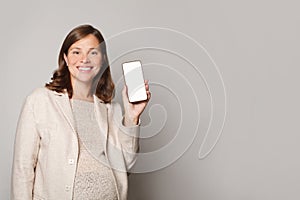 Happy beautiful pregnant woman showing her mobile phone with white empty display and smiling against grey studio wall background