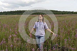 Happy Beautiful smiling blond young woman in a light purple shirt on a meadow of fireweed flowers and looking up at the sky at