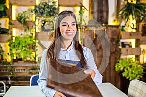 Happy beautiful smiling waitress wearing apron giving a folder menu in a restaurant, looking at camera, standing in cozy