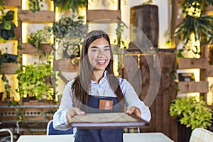 Happy beautiful smiling waitress wearing apron giving a folder menu in a restaurant, looking at camera, standing in cozy