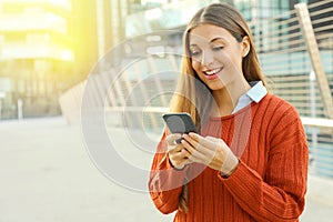 Happy beautiful student woman writing or reading sms messages online on a smart phone while standing in modern city street. Copy